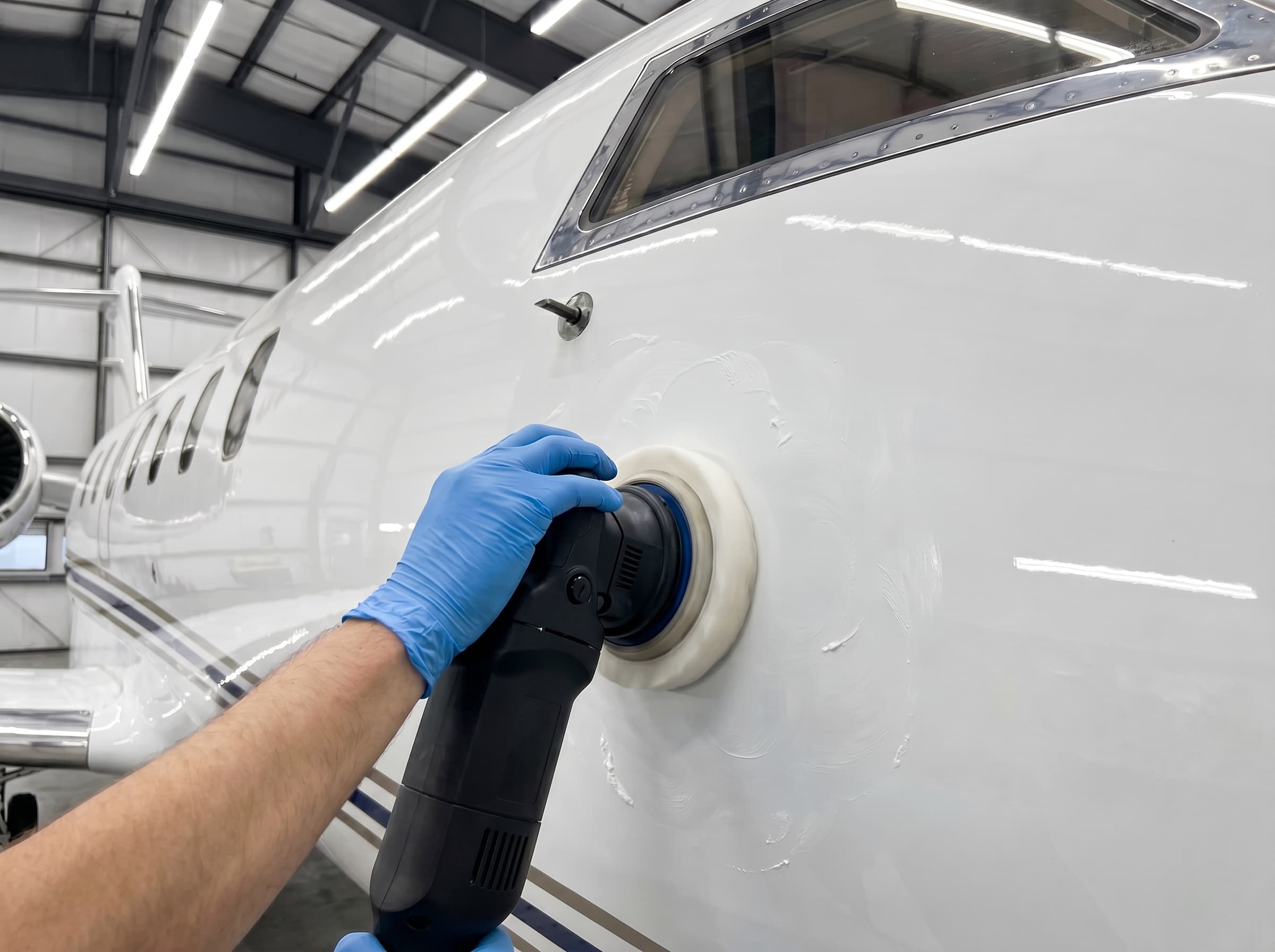Gloved hand operating a dual-action polisher on a white aircraft fuselage panel